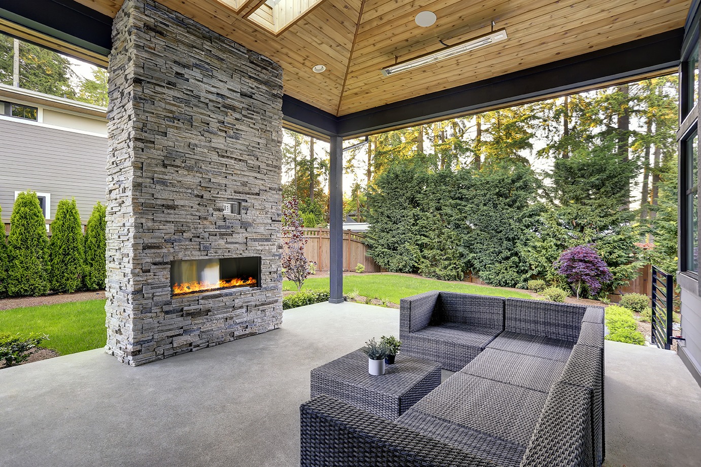 New modern home features a backyard with covered patio accented with stone fireplace, vaulted ceiling with skylights and furnished with gray wicker sofa placed on concrete floor. 