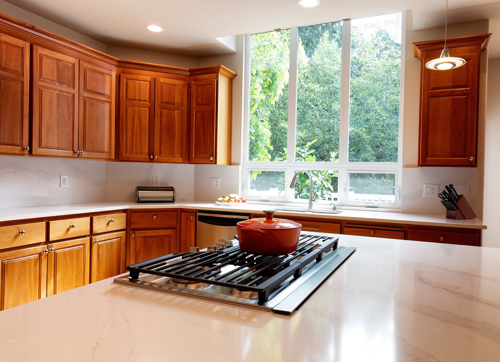 Close up of kitchen stovetop with stone counter top, cherry cabinets and a large window with daylight coming in