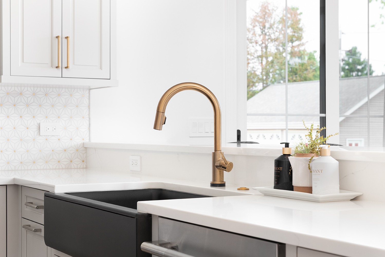 A beautiful sink in a remodeled modern farmhouse kitchen with a gold faucet, black apron or farmhouse sink, white granite, and a tiled backsplash.