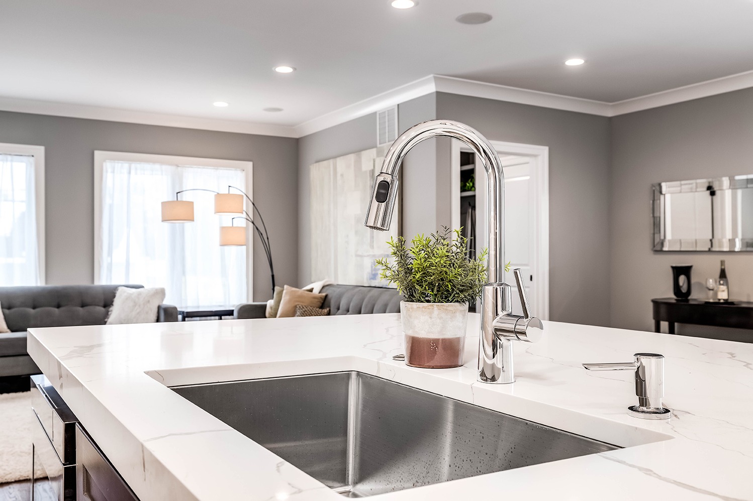 A kitchen sink in a luxurious home looking out towards a living room area. The kitchen island features a white granite and stainless steel tub.