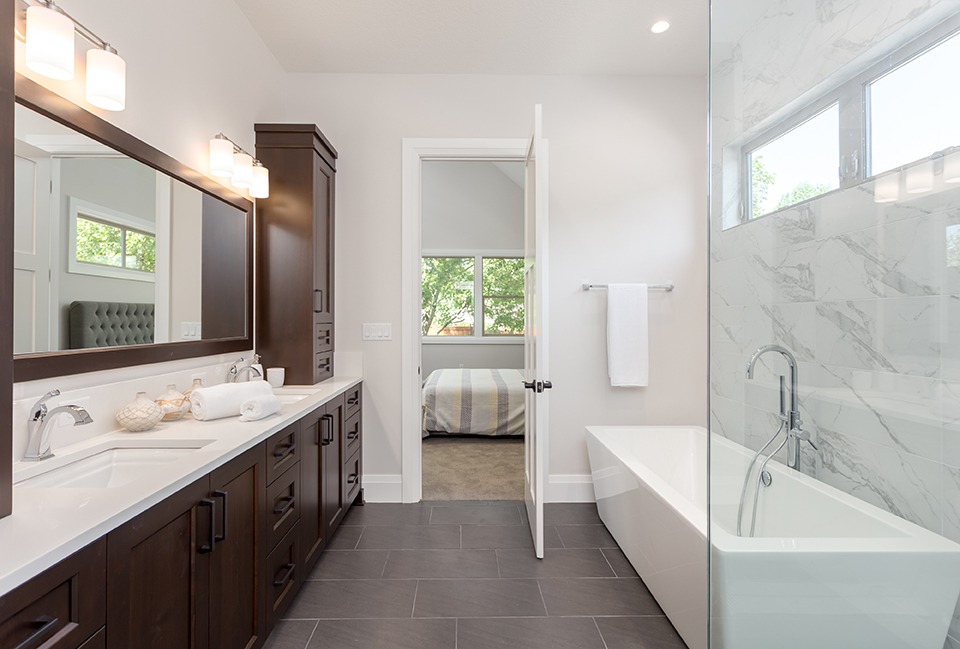 Master bathroom in new luxury home. Tile stretches from floor to ceiling behind bathtub, and partial glass wall hints at luxurious shower. Dark wood cabinetry surrounds double vanity; two sinks.