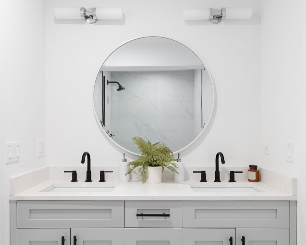 A renovated bathroom with a grey vanity cabinet, circular mirror with a view to a shower, and back faucets.